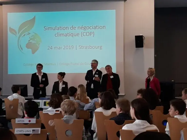 Un groupe d'adultes, parmi lesquels plusieurs professeurs du Jardin des Sciences, se tient devant la classe lors d'un exercice de simulation de négociations sur le climat ; les élèves sont assis et une présentation est projetée sur l'écran derrière eux, ce qui permet aux participants de réécouter les conférences par la suite.