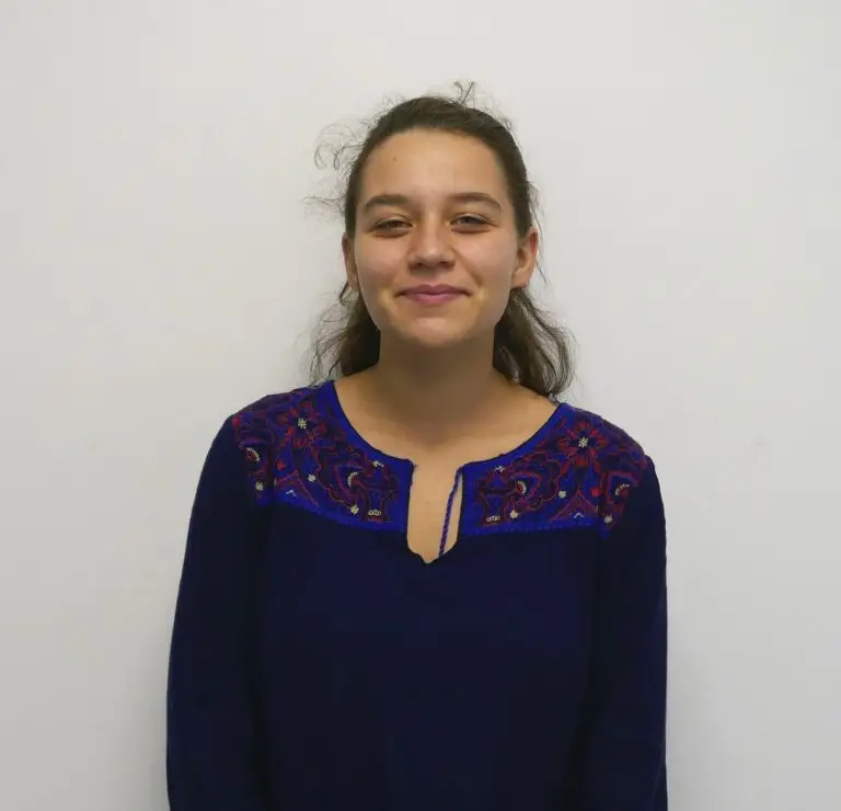 Une jeune femme aux longs cheveux châtains sourit doucement, debout devant un fond blanc uni, vêtue d'un haut bleu marine orné de broderies colorées sur les épaules — une image qui incarne la bonne humeur de Léa Santerre à l'occasion de la rentrée 2021.
