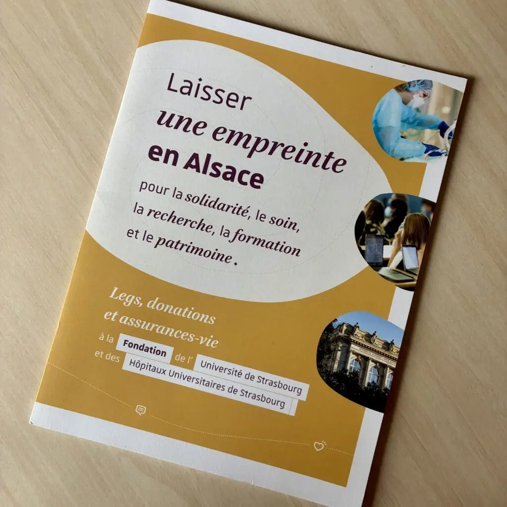 Une brochure française consacrée aux donations en Alsace, illustrée de photos représentant des établissements de santé, des centres de recherche et un bâtiment historique, est posée sur une table en bois clair. Cette brochure présente différentes options, telles que les assurances-vie et les dons, pour soutenir des causes locales.