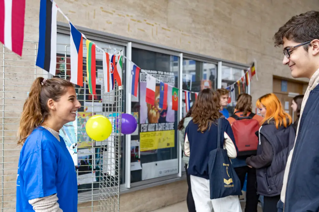 Des étudiants font la queue près d'un stand décoré de drapeaux du monde entier et de ballons. Une jeune femme en chemise bleue sourit à un jeune homme tandis que d'autres attendent à proximité, impatients de savoir comment ils peuvent faire des dons pour soutenir les étudiants étrangers. Des affiches sont visibles sur le mur derrière eux.