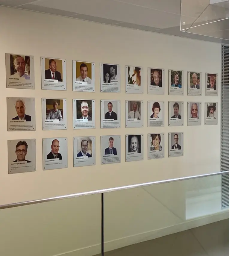 Une exposition murale composée de 20 portraits encadrés et de plaques nominatives représentant diverses personnalités, chacune accompagnée d'une photo et d'une plaque descriptive, est disposée sur quatre rangées à l'intérieur d'un bâtiment moderne, rappelant le « mur des donateurs » dévoilé lors de l'inauguration à Sciences Po Strasbourg.