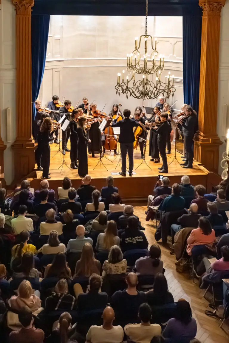 L'Orchestre universitaire de Strasbourg se produit sur scène devant un public dans une salle de concert aux murs lambrissés, aux rideaux bleus et ornée d'un lustre, marquant ainsi une année exceptionnelle tant pour les musiciens que pour leurs auditeurs.