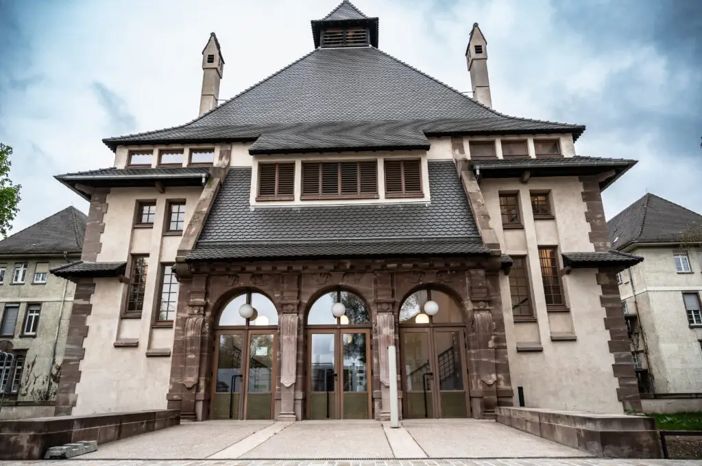 Un vaste édifice symétrique, avec un toit sombre et en pente raide, des portes voûtées et des colonnes en pierre, se découpant sur un ciel nuageux : « La Fondation » se dresse, imposante, à l'horizon.