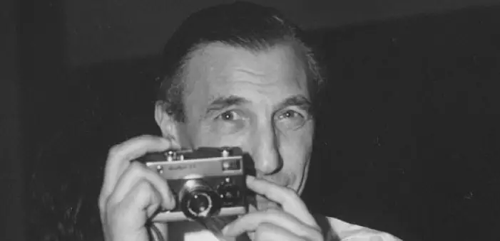 Un homme aux cheveux lissés en arrière tient un appareil photo ancien devant son visage, le regard tourné directement vers l'objectif. L'image est en noir et blanc.