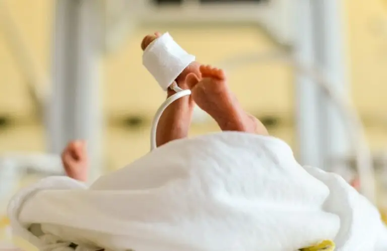 Les jambes d’un bébé prématuré, équipées d’un capteur médical, allongées sur un lit d’hôpital recouvert d’une couverture blanche, avec du matériel médical visible à l’arrière-plan : une image poignante qui rappelle à quel point les dons peuvent contribuer à financer des soins néonatals vitaux.