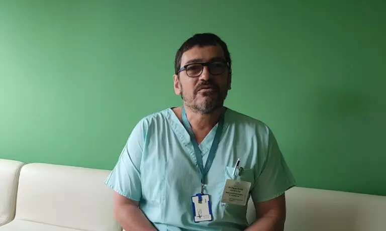 Un homme vêtu d'une blouse médicale vert clair et portant un badge d'identification, probablement impliqué dans le projet NIDCAP, est assis sur un canapé blanc devant un mur vert uni.