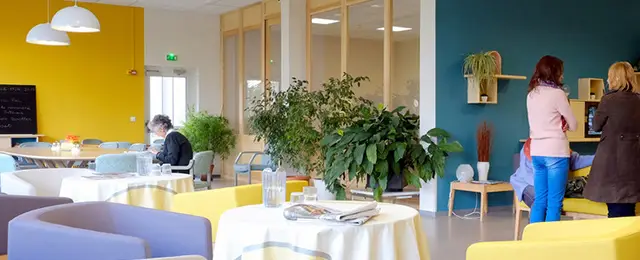 Un espace de détente lumineux et moderne, aux murs jaunes et bleu sarcelle — issu d’une récente rénovation de l’accueil —, est agrémenté de tables, de chaises et de plantes disposées un peu partout, ainsi que de trois personnes : l’une assise en train de lire et les deux autres debout en train de discuter. Cet espace « Bon Séjour », très accueillant, améliore la prise en charge des patients en leur offrant une atmosphère confortable propice à la détente et à la conversation.