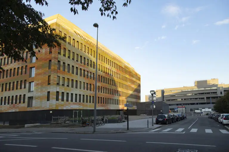 Un immeuble moderne de plusieurs étages, à la façade jaune et orange, qui abrite l'Institut de génétique médicale d'Alsace (IGMA), se dresse au bord d'une rue calme où sont garées des voitures et des vélos, sous un ciel dégagé.