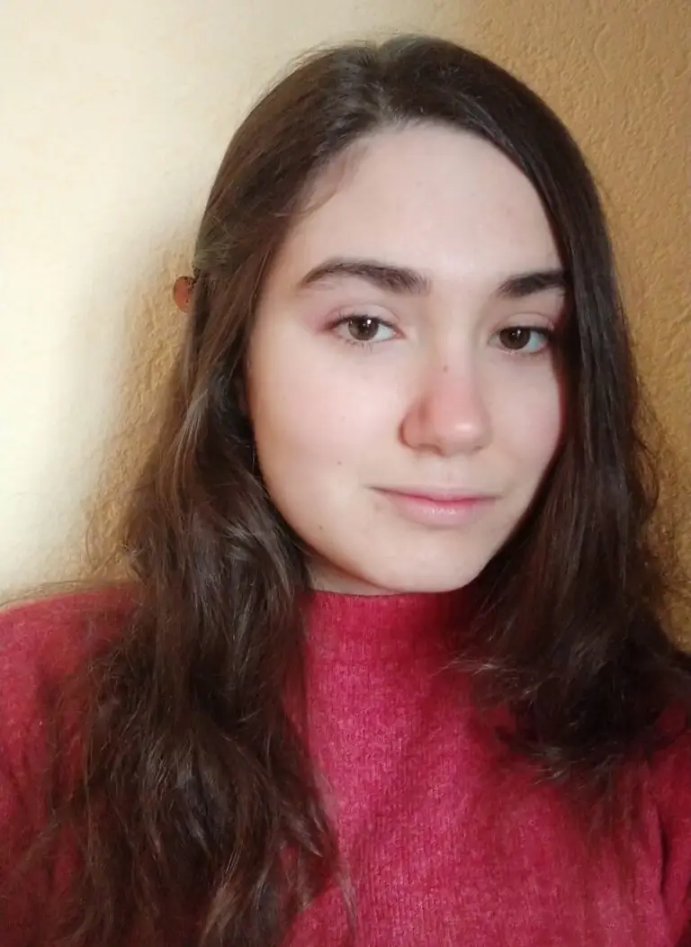 Un jeune aux longs cheveux châtains, vêtu d'un pull rouge, se tient debout devant un mur beige, le regard tourné vers la caméra avec une expression neutre, prêt à partager son témoignage sur l'obtention d'une bourse grâce à la générosité de donateurs.