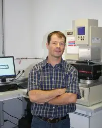 Un homme vêtu d'une chemise à carreaux se tient debout, les bras croisés, dans un laboratoire, à côté d'appareils scientifiques et d'un ordinateur.
