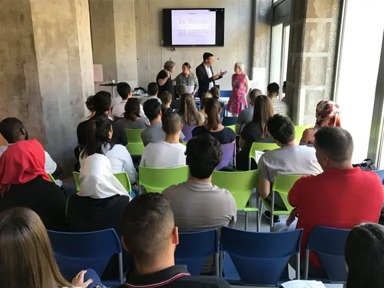 Un groupe de personnes est assis et assiste à une présentation au cours de laquelle plusieurs intervenants se tiennent à l'avant de la salle, près d'un écran sur lequel s'affiche du texte.