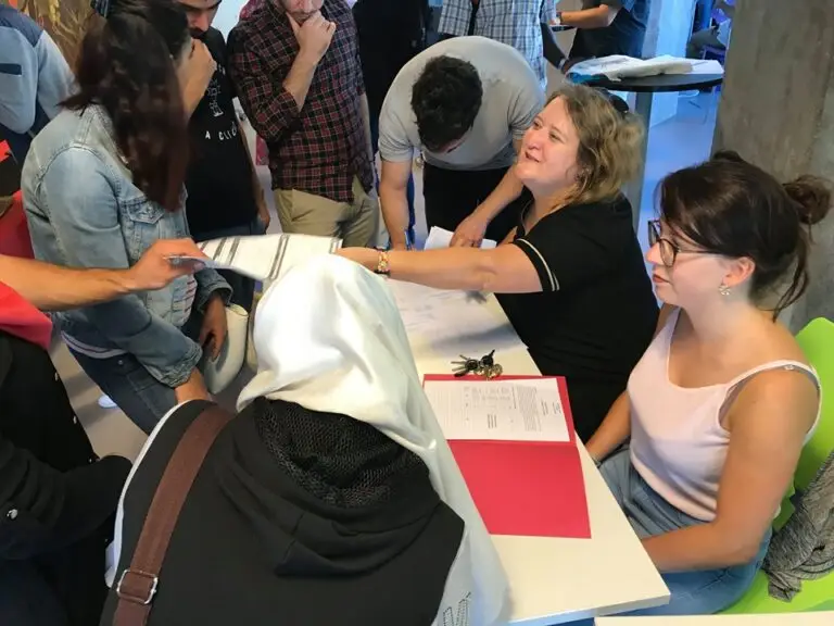 Un groupe de personnes se rassemble autour d'une table recouverte de papiers, tandis que deux femmes assises les aident à s'installer à l'intérieur, illustrant ainsi les valeurs humanistes défendues par l'Université de Strasbourg dans son soutien aux étudiants réfugiés.