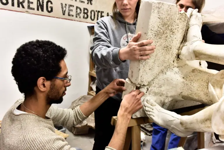 Trois personnes travaillent ensemble à la restauration ou à l'assemblage d'une grande sculpture blanche représentant des jambes humaines, dans ce qui semble être un atelier d'art, évoquant le processus minutieux de rénovation des œuvres souvent mené dans des lieux tels que le musée Adolf Michaelis.