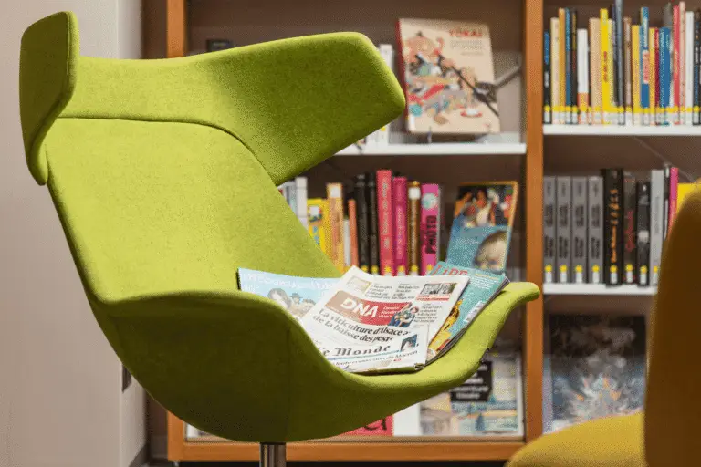 Une chaise moderne de couleur verte, sur laquelle sont posés plusieurs journaux, est placée devant une bibliothèque remplie de livres et de magazines, créant ainsi un espace idéal pour lire le présent tout en étant entouré d'ouvrages qui préservent le passé.