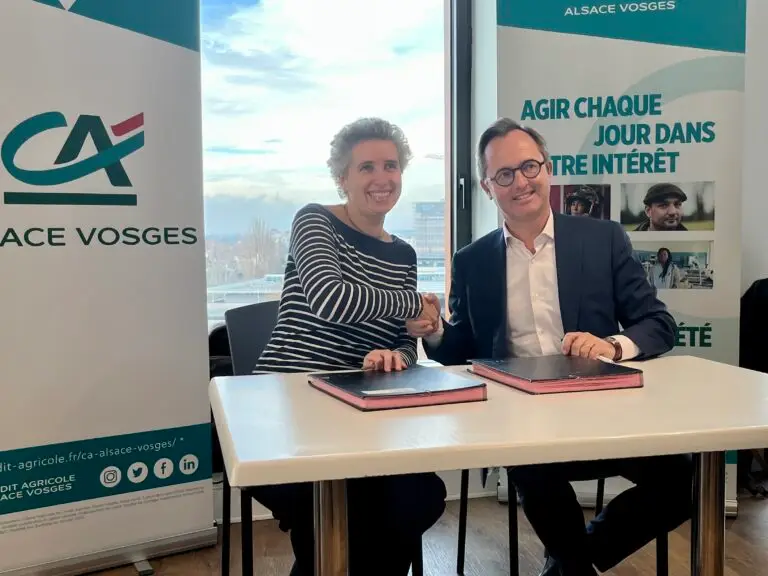 Deux personnes sont assises à une table, se serrant la main et souriant, avec des documents devant elles et des bannières promotionnelles en arrière-plan — mettant en avant un partenariat de mécénat entre le Crédit Agricole Alsace Vosges et l'Université de Strasbourg.