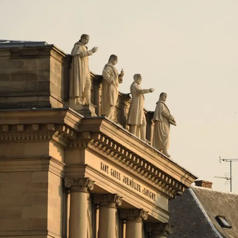 Quatre statues de pierre, chacune se dressant fièrement au sommet d’un édifice classique à colonnes, confèrent à la scène une atmosphère de force sereine. Les noms de Kant, Gauss, Dollmüller et Savigny sont gravés en relief sous leurs pieds. À proximité, on aperçoit un toit et une antenne, témoins silencieux de l’héritage préservé ici grâce au soutien et aux dons de plusieurs générations.