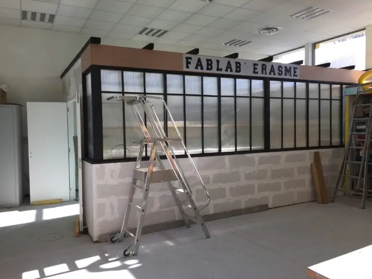 Un escabeau métallique se dresse devant une pièce en cours d'aménagement portant l'inscription « FABLAB ERASME », dont les parois en verre et en panneaux s'intègrent dans un espace de travail baigné de lumière, reflétant l'esprit d'innovation des FabLabs que l'on retrouve dans des environnements éducatifs tels que la Maison pour la Science en Alsace ou un collège moderne.