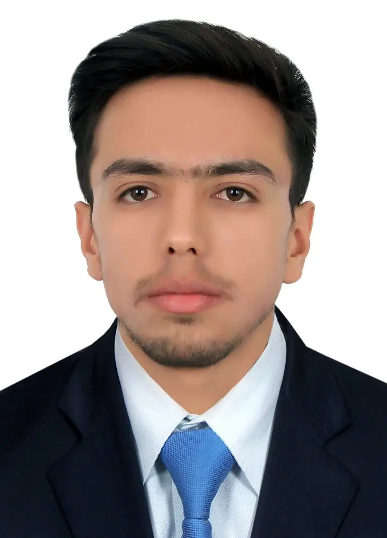 Un homme aux cheveux courts et foncés, portant une barbe, étudiant réfugié originaire d'Afghanistan, vêtu d'un costume sombre, d'une chemise blanche et d'une cravate bleue, fait face à la caméra devant un fond blanc uni à Strasbourg.
