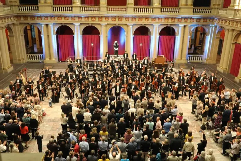 Un orchestre symphonique se tient sur la scène d'une salle de concert sous les applaudissements du public, rappelant le célèbre World Doctors Orchestra qui avait donné à Strasbourg un concert inspiré par les soignants. Les musiciens tiennent leurs instruments, tandis que des rideaux rouges et des colonnes encadrent la scène.
