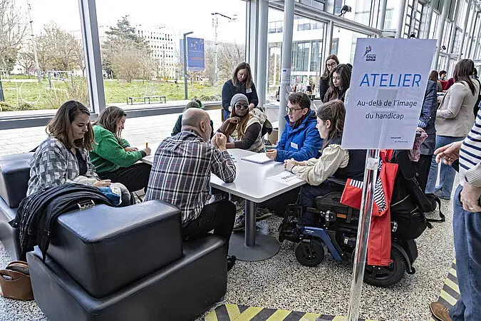 Un groupe de personnes, parmi lesquelles une personne en fauteuil roulant, est assis autour d'une table dans un atelier d'Emploi'Ability baptisé « Atelier », situé dans un espace intérieur lumineux doté de grandes baies vitrées, ce qui témoigne de leur engagement en faveur de l'inclusion et de l'égalité d'accès aux opportunités d'emploi.