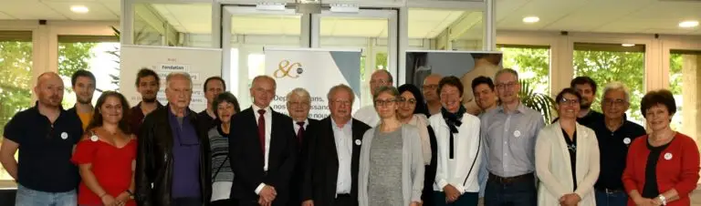 Un groupe d'adultes, hommes et femmes, se tient à l'intérieur devant de grandes baies vitrées et des banderoles, posant ensemble pour une photo de groupe.