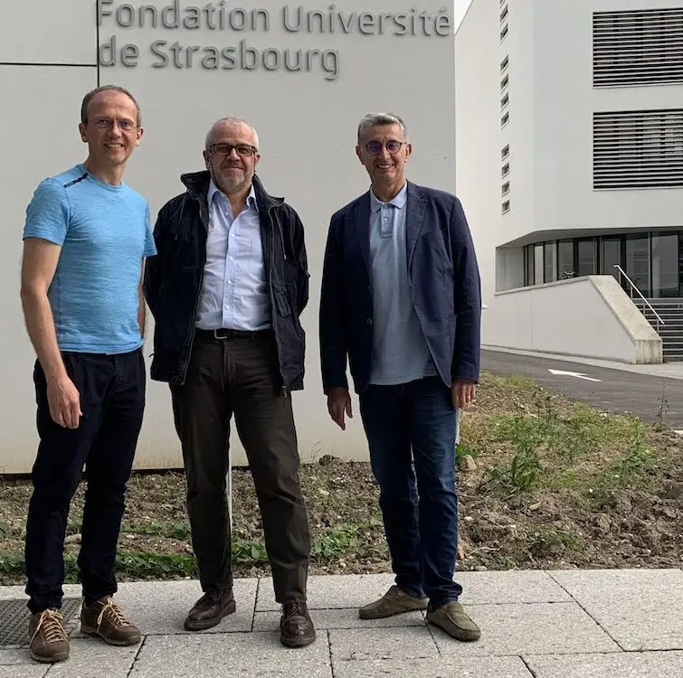 Trois hommes se tiennent devant le bâtiment de la Fondation Université de Strasbourg, posant pour une photo sur une allée pavée près d'une façade blanche moderne ; leur conversation est subtilement influencée par les récentes discussions concernant l'épidémie de Covid-19.