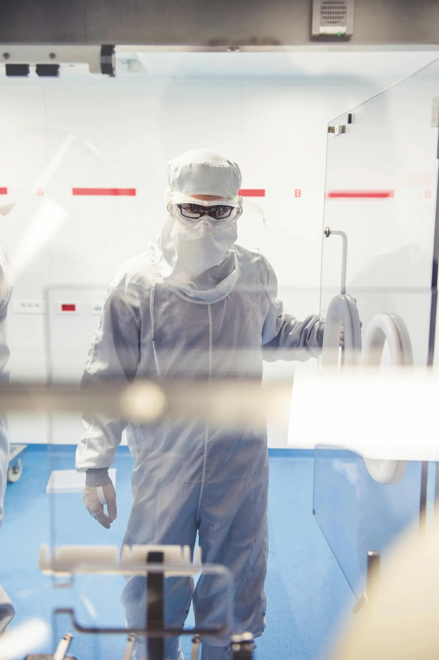 Une personne vêtue d’une combinaison de protection intégrale et de lunettes de protection se tient dans un laboratoire stérile, une main posée sur une porte — illustrant ainsi les normes de sécurité rigoureuses qui sont au cœur du « projet majeur » mené à l’usine-école EASE de l’Université de Strasbourg.