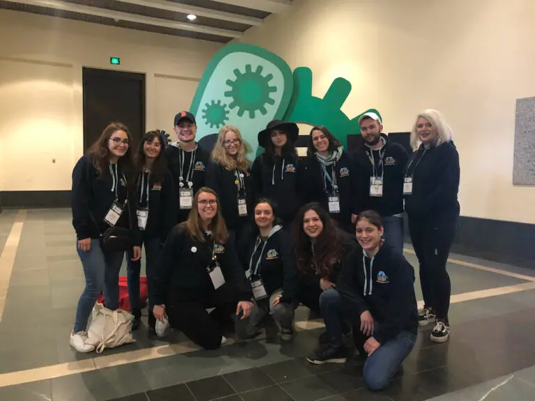 Un groupe de treize personnes, vêtues de sweats à capuche assortis et portant des badges de conférence, posent à l'intérieur devant une sculpture verte en forme d'engrenage, représentant fièrement leur équipe lors du concours international iGEM.
