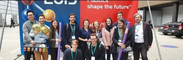 Un groupe de treize personnes, dont des étudiants de l’ECPM en voyage d’étude à K2019, posent en extérieur devant des bannières ornées d’illustrations à thème scientifique et portant la mention « Les plastiques façonnent l’avenir ». Certains tiennent des accessoires et tous portent leur badge de la conférence.