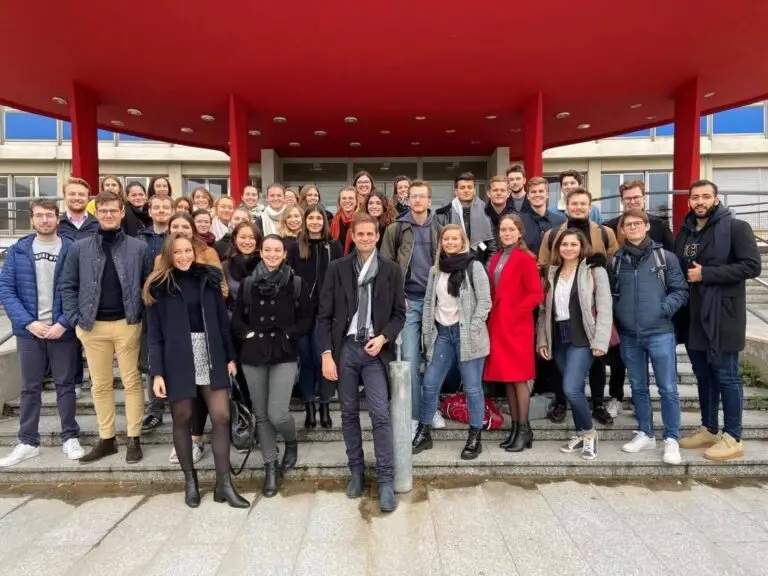 Un groupe d’une quarantaine de personnes, participant au voyage d’étude du Master II en droit bancaire et financier, pose pour une photo sur les marches situées devant un bâtiment aux colonnes rouges et doté d’un auvent rouge incurvé.