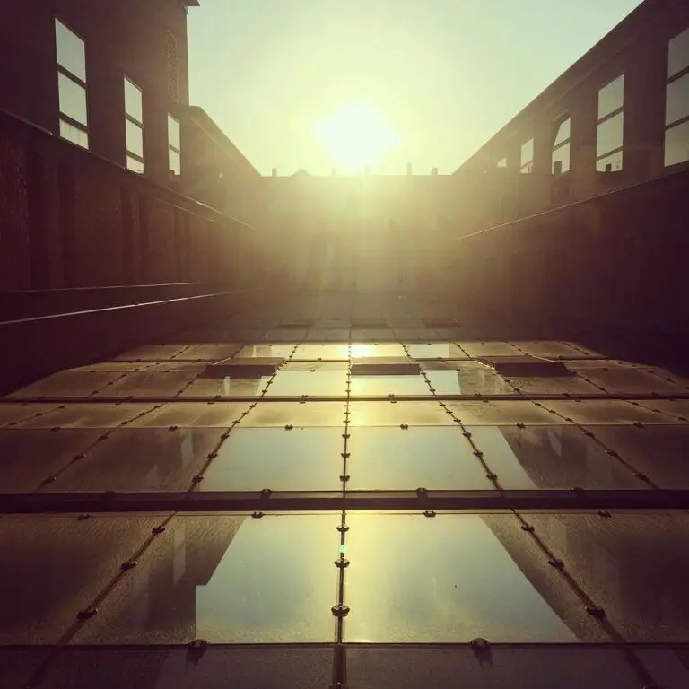 La lumière du soleil filtre entre deux bâtiments en briques, se reflétant sur une passerelle ou un toit en verre, tandis que le soleil est bas dans le ciel : une scène qui illustre à merveille la synergie et la créativité fondamentale qui naissent lorsque les éléments architecturaux s’engagent dans une subtile « coopétition ».