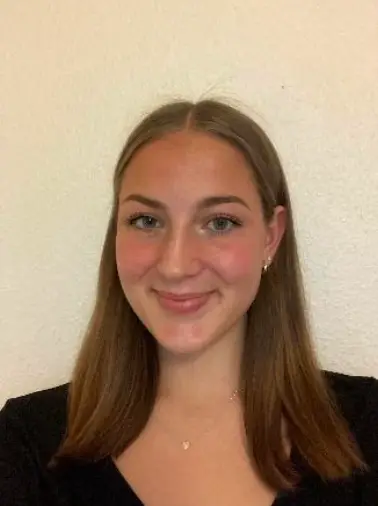 Mégane Lett, une jeune femme aux cheveux bruns raides et au teint clair, sourit à la caméra, debout devant un mur uni de couleur claire. Étudiante boursière, elle porte un haut noir et de petites boucles d'oreilles, prête pour son entretien.