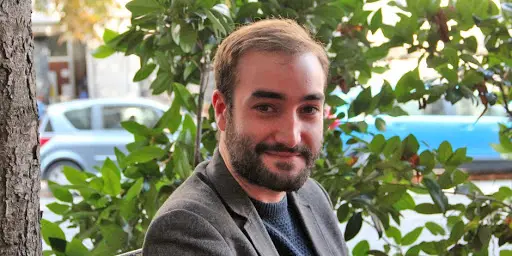 Un homme barbu, vêtu d'un blazer gris et d'une chemise foncée, est assis en plein air près de buissons verts, avec des voitures garées et une rue en arrière-plan.