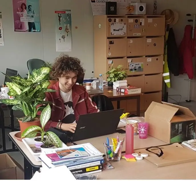 Une personne aux cheveux bouclés est assise devant un bureau encombré, travaillant sur un ordinateur portable, entourée de plantes, de fournitures de bureau, de livres et de piles de cartons en arrière-plan — plongée dans des projets innovants pour Biotech-Lab.
